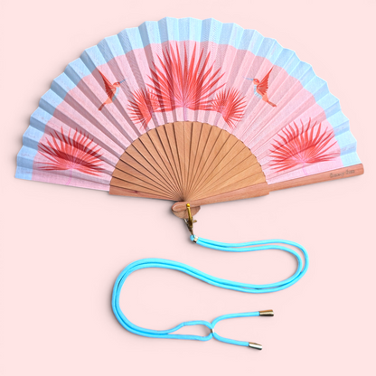 LE BAZAR INOUI BIRDY Handfächer mit Kolibri-Motiv, rosa und hellblauem Stoff auf einem Holzrahmen mit blauem Trageband.