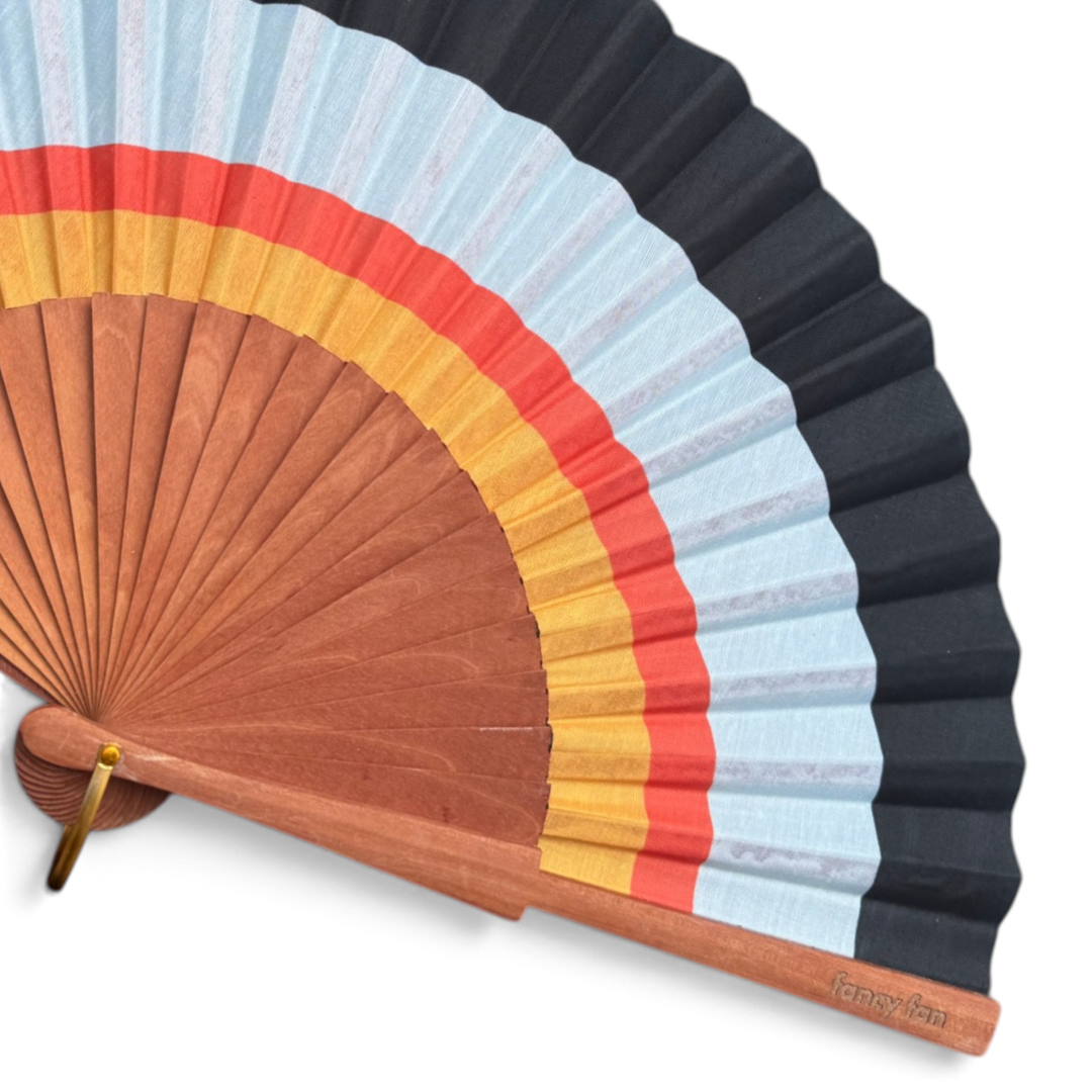 LE BAZAR INOUI HORIZON Handfächer aus Holz und Stoff mit farbenfrohem Streifenmuster in Schwarz, Rot, Gelb und Blau.